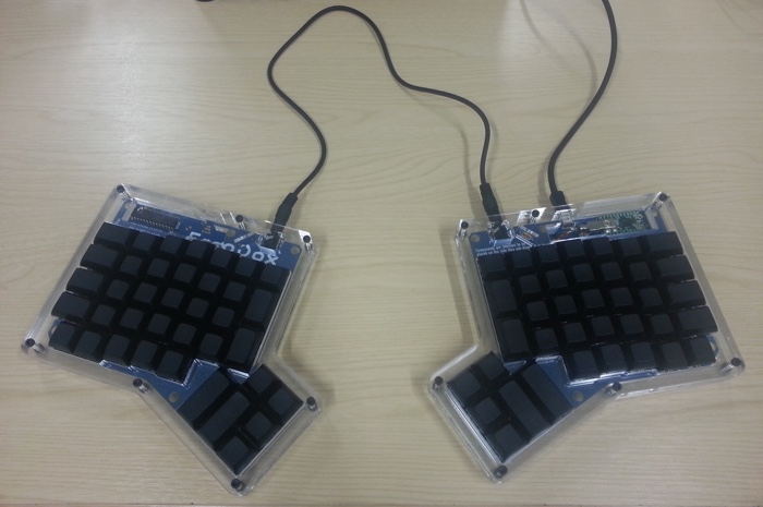 
   My completed keyboard fully-assembled and plugged in on a desk.
   It's a split keyboard with the two pieces connected by a cable.
   The keycaps are unlabeled and black.