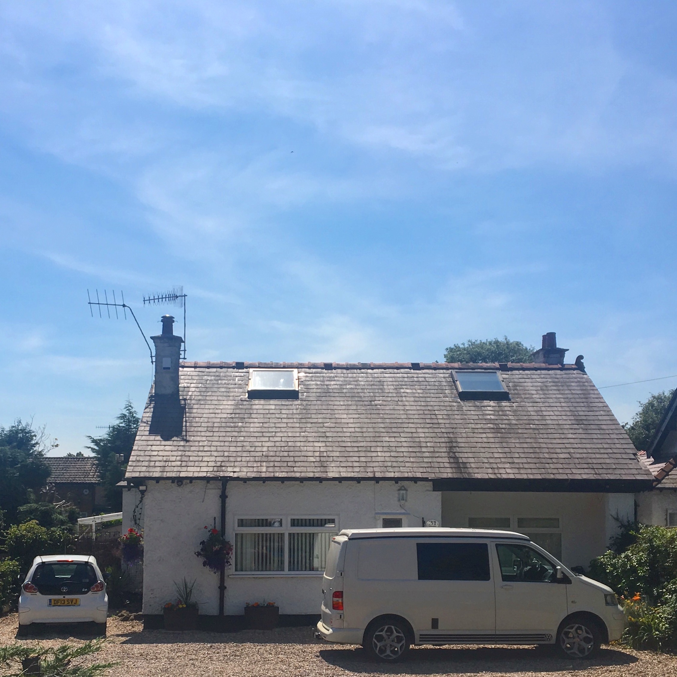 White bungalow with a grey roof on a clear sunny day. A white car and van are parked outside on a gravel drive.