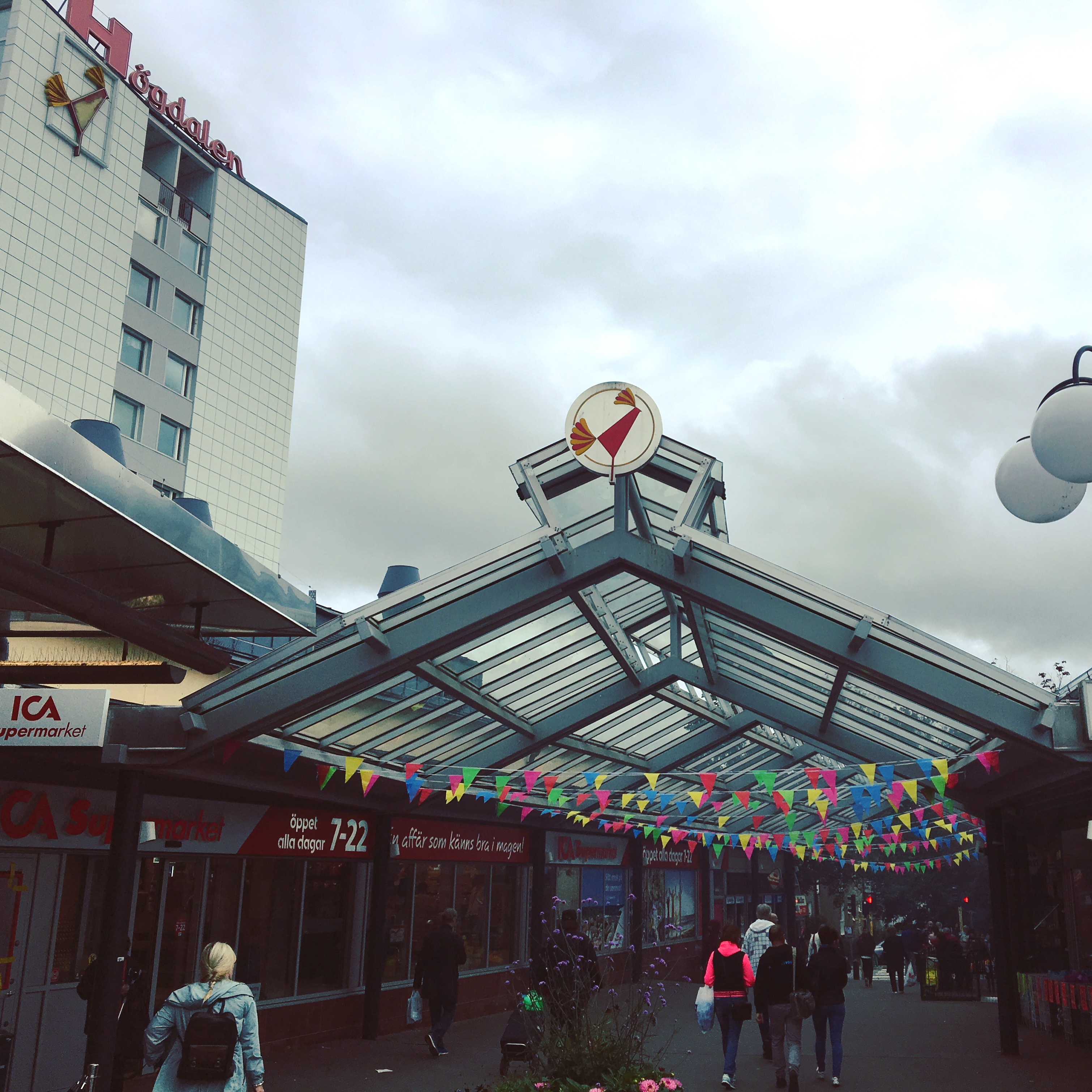 Busy street scene with people coming and going, multicolored bunting hanging above, as well as a circular white sign with a red rooster symbol. The same symbol is emblazoned in a 3 meter tall sign on a tall building nearby, underneath a large sign saying “Högdalen”.