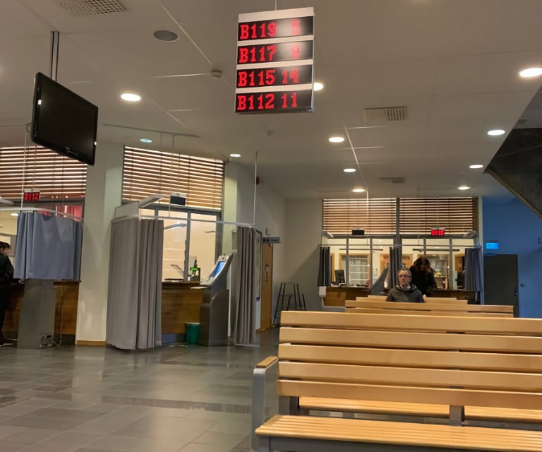 Waiting room with fluorescent lights and an LED sign showing current queue positions. Wooden benches in the foreground. Behind them are several booths with curtains around them for taking flash photos inside.