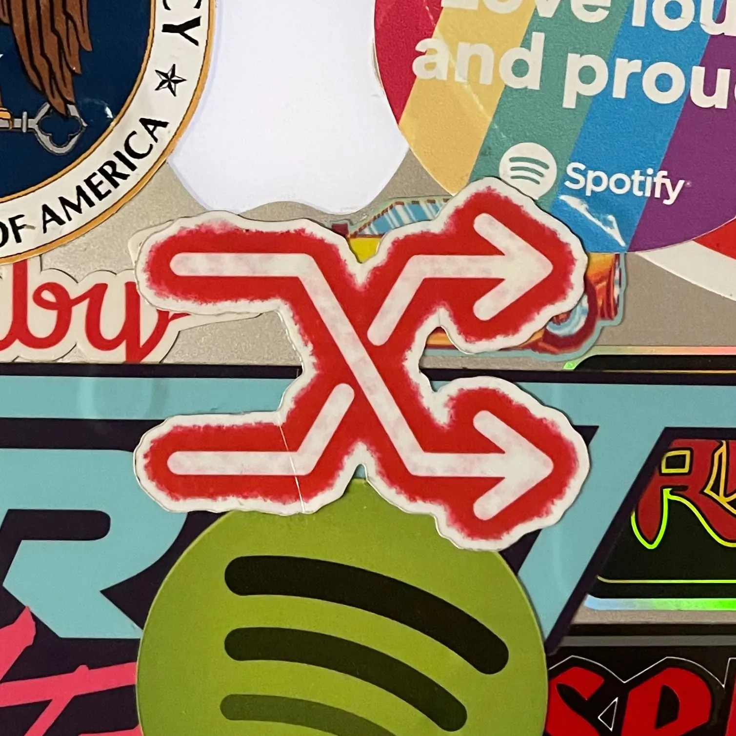 A shuffle symbol with a white foreground and a red spraypaint outline is seen affixed as a sticker to a laptop next to other stickers.