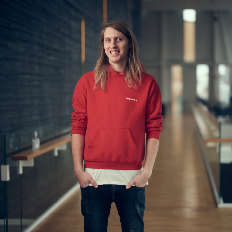 Person with long blonde hair in a bright red hoodie stands in modern-looking hotel lobby.