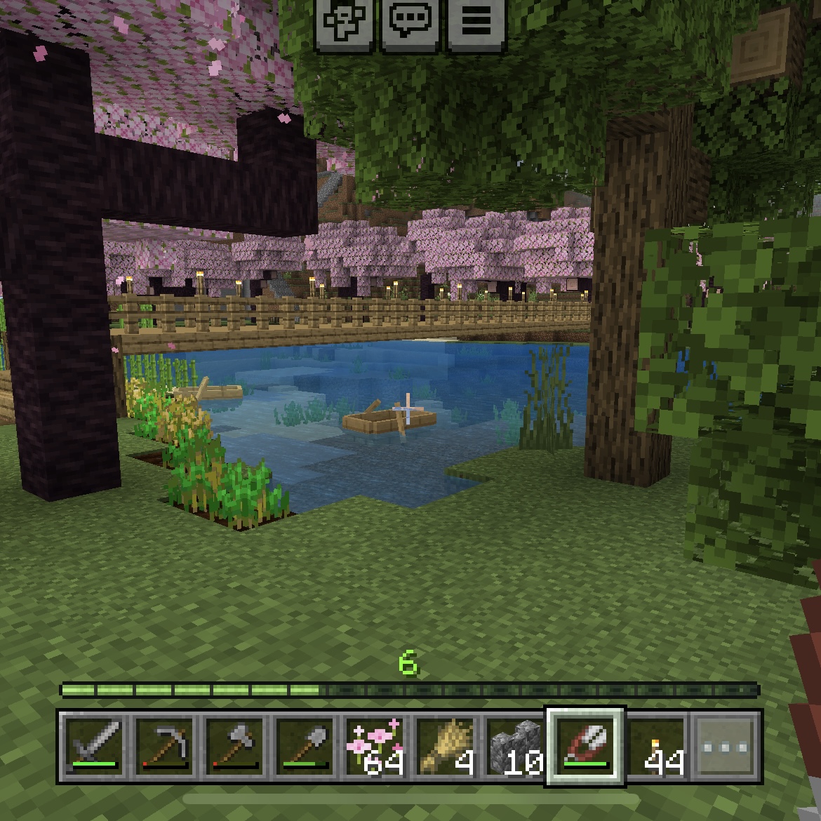 Minecraft screenshow featuring a pixelated wooden boat in some shallow water in the shade of a cherry tree and an oak tree with a wooden bridge in the background.