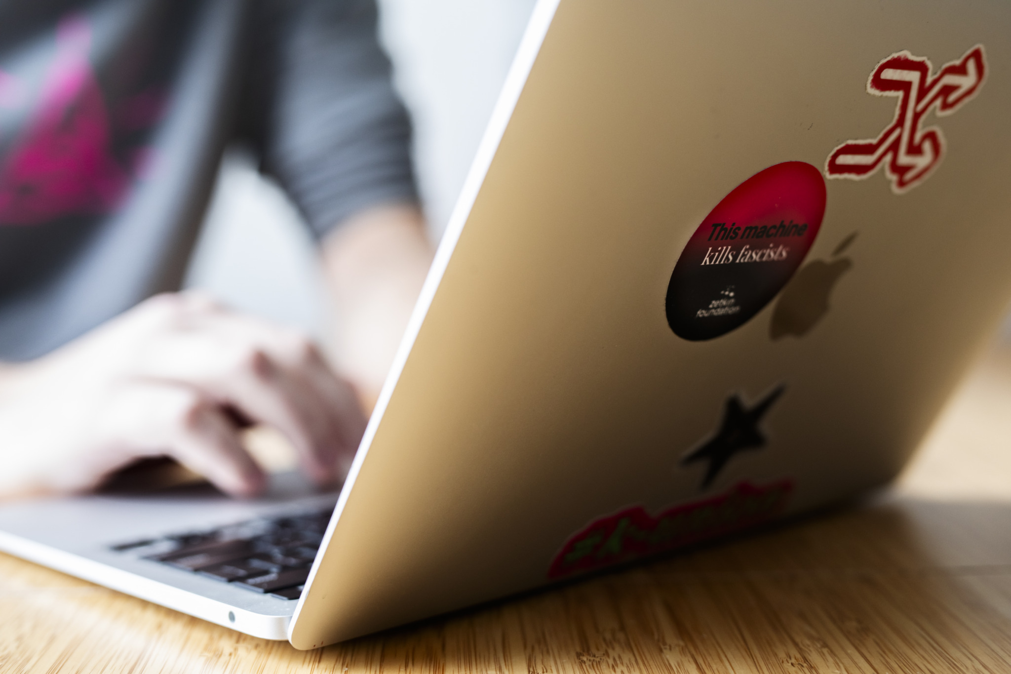 Laptop on table in the foreground, person in a grey hoodie typing in the background. A sticker reading 'This machine kills fascists' is visible on the laptop.