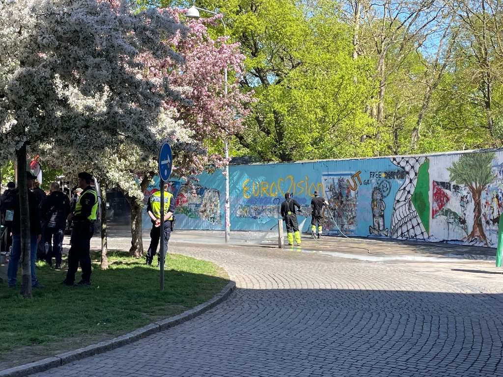 Armed police guarding a clean up crew steam cleaning the area around a graffiti mural featuring a Eurovision logo and a Palestine flag.