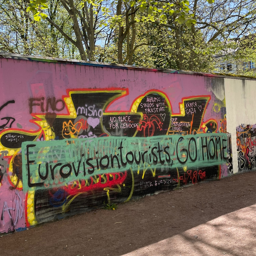 Graffiti mural reading 'Eurovision tourists GO HOME' with messages of solidarity for Gaza in smaller text around it.