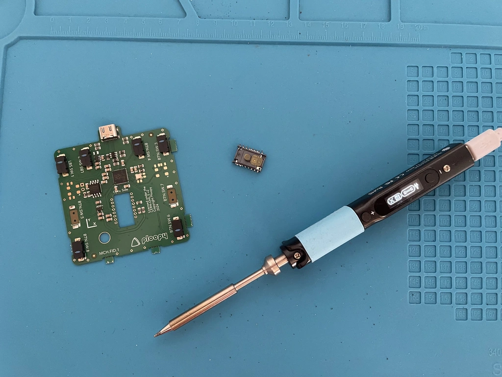 A blue soldering mat with a PCB, a microchip, and a soldering iron.